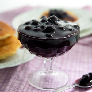 Fancy goblet glass with Very Berry Syrup inside and pancakes in the background on a light pink tablecloth