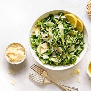 Arugula Salad with Lemon Balsamic in a white bowl with bowls of pine nuts, parmesan cheese and dressing on the side