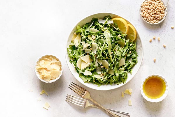 Arugula Salad with Lemon Balsamic in a white bowl with bowls of pine nuts, parmesan cheese and dressing on the side