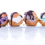 Row of four Gluten-Free Profiteroles stuffed with different colors of ice cream and drizzled with chocolate sauce on a white background