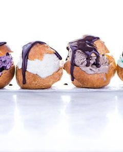 Row of four Gluten-Free Profiteroles stuffed with different colors of ice cream and drizzled with chocolate sauce on a white background