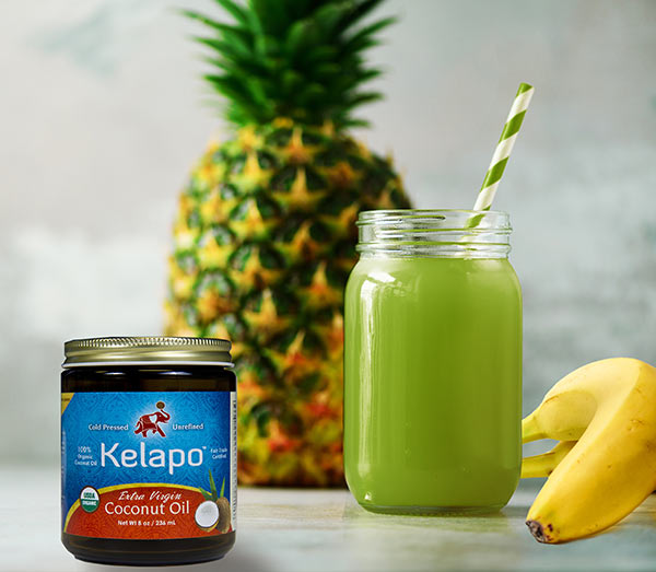Jar of Kelapo coconut oil in front of a green smoothie with banana and whole pineapple in the background