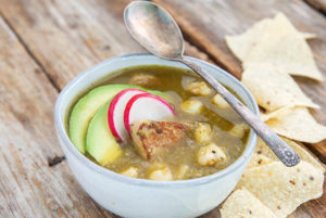 Pork Pozole Verde in a white bowl on a wooden background with tortilla chips on the side