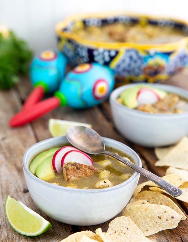Pork Pozole Verde in white bowls on a wooden table with tortilla chips and limes in the background