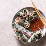 Sesame Kale Spring Rolls in a white and beige bowl with sauce on the side and wooden chopsticks