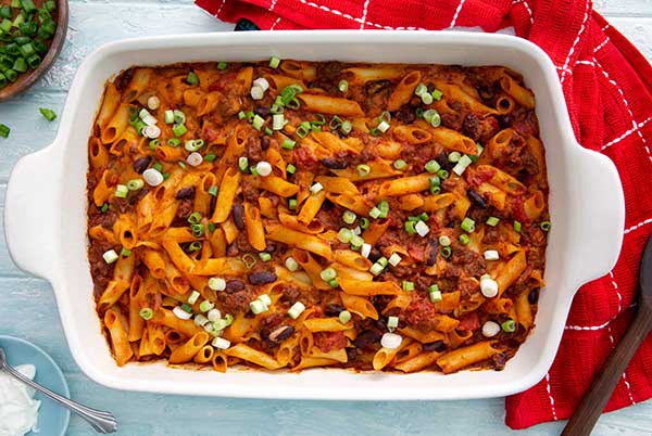 Overhead view of Plant-Based Chili Sin Carne Pasta Bake in a white baking dish with red kitchen towel underneath on a pale blue background