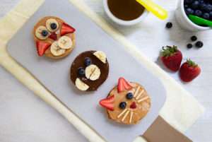 Overhead view of Animal Face Bagels on a ceramic board with bowls of blueberries and chocolate hazelnut spread
