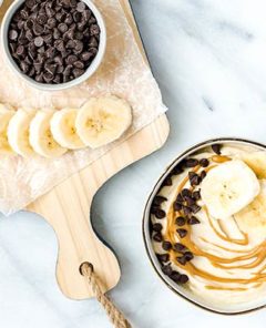 Overhead view of Banana Nice Cream in a bowl topped with sliced banana and chocolate chips and peanut butter with a cutting board next to it with more of those toppings spread out