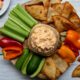 Overhead view of Chili Pimento Cheese in a jar surrounded by tortilla chips and fresh veggies on a round wooden board