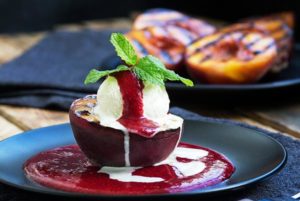 Grilled Peach Melbas with raspberry sauce and mint leaves on top of a black plate with grilled peaches in the background