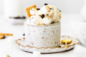 No-Churn S'mores Ice Cream in a white and brown speckled cup with a gold spoon next to it and mini marshmallows scattered