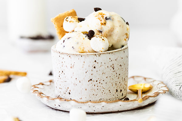 No-Churn S'mores Ice Cream in a white and brown speckled cup with a gold spoon next to it and mini marshmallows scattered