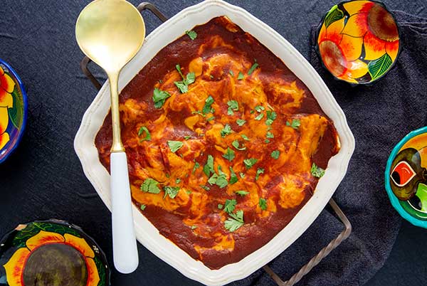 Overhead view of Plant-Based Enchilada Lasagna in a square white casserole dish with gold and white spoon on a dark gray table
