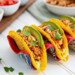 Salsa Chicken in hard corn taco shells in a red and blue ceramic taco holder on a white table with lime wedges and tomatoes in the background