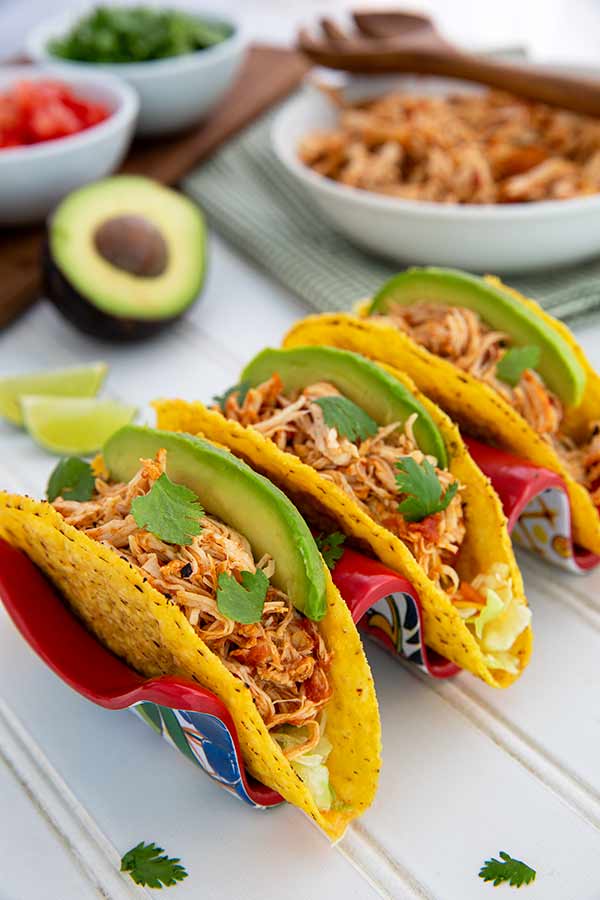Salsa Chicken in hard corn taco shells in a red and blue ceramic taco holder on a white table with avocado and tomatoes in the background