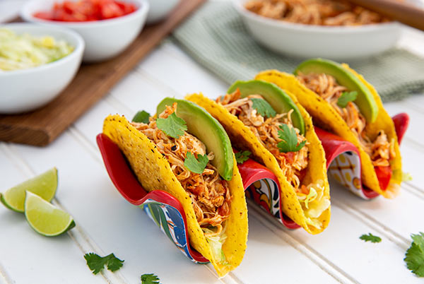 Salsa Chicken in hard corn taco shells in a red and blue ceramic taco holder on a white table with lime wedges and tomatoes in the background