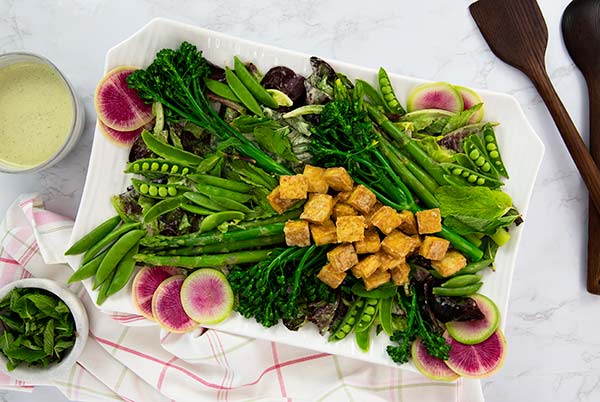 Overhead view of Spring Salad with Crispy Tofu Croutons on a white rectangle serving platter with ramekins of dressing and green onion on the side and wooden serving utensils