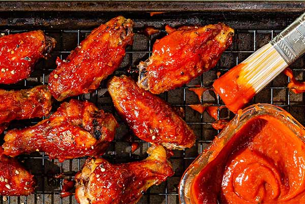 Overhead view of Sticky Mango Chicken Wings on a wire rack on a baking sheet with glass bowl of sauce and sauce brush