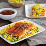 Barbecue Salmon with Corn Salsa on two rectangular white plates with a bowl of sauce and bowl of salsa next to them on a dark wood table