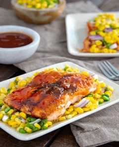 Barbecue Salmon with Corn Salsa on two rectangular white plates with a bowl of sauce and bowl of salsa next to them on a dark wood table