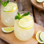 Healthy Lemon-Lime Fizz drink in stemless wine glasses garnished with lemon and lime slices and mint on a wooden board on a darker wood table