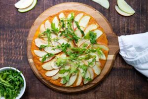 Overhead view of Plant-Based Apple and Cheese Flatbread on a wooden pizza board on a darker wood table with pizza cutter next to it