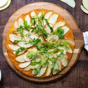 Overhead view of Plant-Based Apple and Cheese Flatbread on a wooden pizza board on a darker wood table with pizza cutter next to it