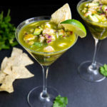 Aguachile Ceviche in two martini glasses topped with a lime wedge and tortilla chip on a black table with a bunch of cilantro and tortilla chips in the background