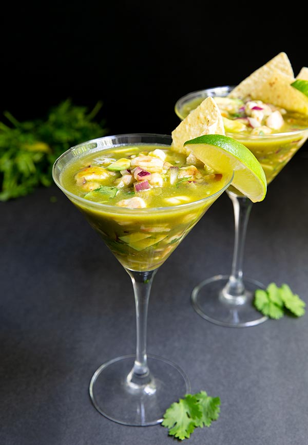 Aguachile Ceviche in two martini glasses topped with a lime wedge and tortilla chip on a black table with black background and bunch of cilantro in the background