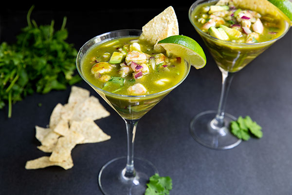 Aguachile Ceviche in two martini glasses topped with a lime wedge and tortilla chip on a black table with a bunch of cilantro and tortilla chips in the background