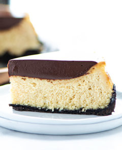 Closeup of a slice of Baileys Cheesecake on a white plate on a white background with the remaining cheesecake in the back