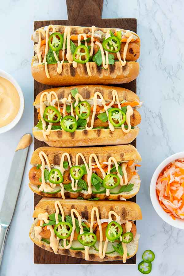 Overhead view of Banh Mi Hot Dogs drizzled with sriracha mayo on a wooden board on a white marble table
