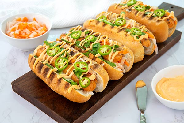 Closeup of Banh Mi Hot Dogs drizzled with sriracha mayo on a wooden board on white marble counter