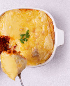 Overhead view of Cheesy Potato Lentil Casserole in a white casserole with a person's hand holding it and taking a spoonful out of the casserole on a white background
