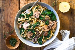 Overhead view of Creamy Mushroom Pasta in a white bowl on a rustic wood table with fresh herbs and lemon around the bowl