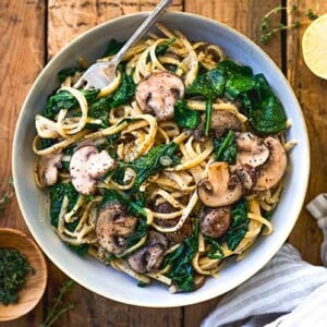 Overhead view of Creamy Mushroom Pasta in a white bowl on a rustic wood table with fresh herbs and lemon around the bowl