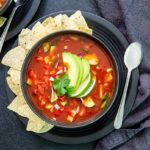 Overhead view of Gazpacho in a black bowl on a black plate with tortilla chips along the side and topped with avocado slices on a black table
