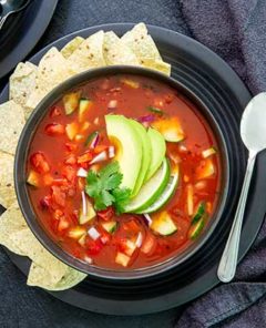 Overhead view of Gazpacho in a black bowl on a black plate with tortilla chips along the side and topped with avocado slices on a black table