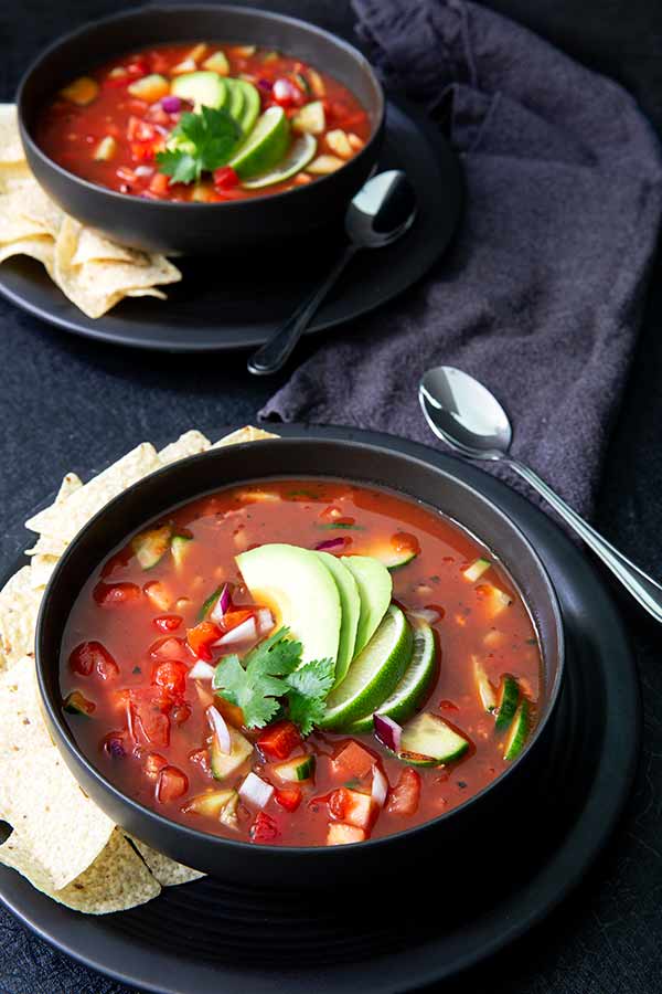 Gazpacho in two black bowls on black plates with tortilla chips on the side and topped with avocado and lime slices on a black tablecloth