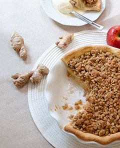 Overhead view of Ginger Apple Pie with fresh knobs of ginger and red apples next to it on a white placement on an off-white table