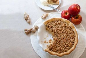 Overhead view of Ginger Apple Pie with fresh knobs of ginger and red apples next to it on a white placement on an off-white table