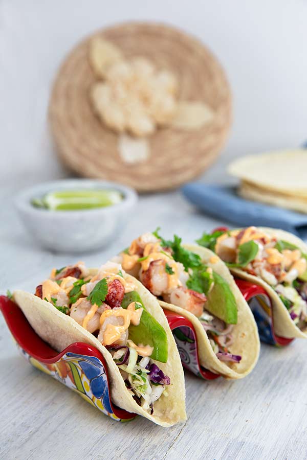 Lobster Tacos with Cilantro Slaw set in a taco holder that has a Mexican art pattern on it on a light gray wood table
