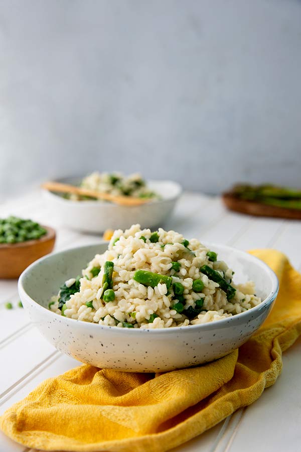 Pea and Asparagus Risotto in a white bowl with mustard yellow napkin underneath on a white table with gray background and bowls of peas and asparagus in the background