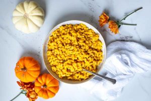 Overhead view of Pumpkin Mac and Cheese in a white bowl with mini white and orange pumpkins along the side on a white marble table