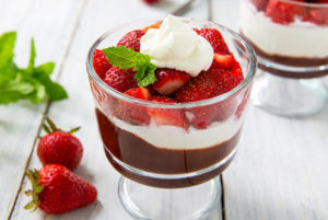 Closeup of Chocolate Strawberry Parfait layered in a clear glass with whole strawberries and mint leaves next to it on a white wood table