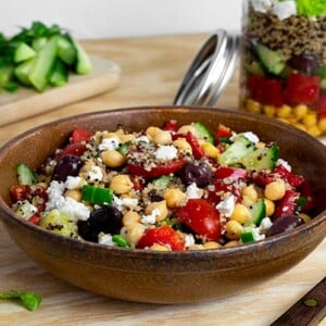 Mediterranean Salad-in-a-Jar in the background with a bowl of the salad in the foreground on a light wood table with salad ingredients in the background