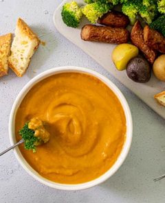 Overhead view of Plant-Based Cheesy Fondue in a white bowl with a hand dipping a broccoli floret into the fondue with a metal skewer and a board of veggies and bread on the side
