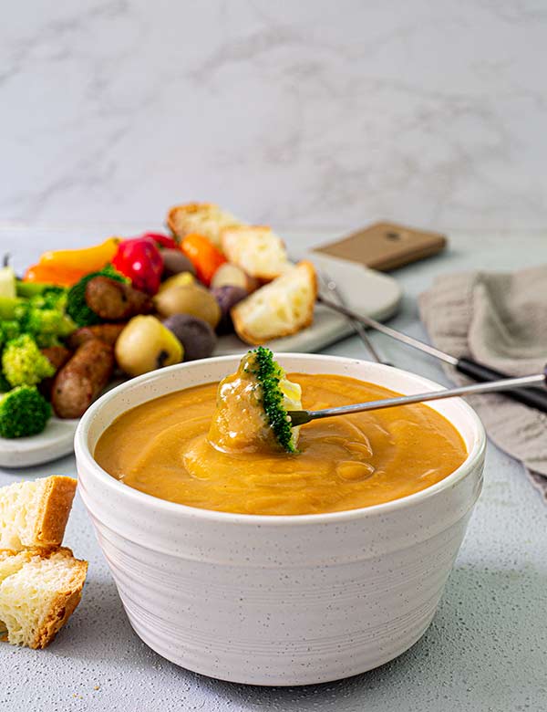 Plant-Based Cheesy Fondue in a white bowl with a skewer dipping a broccoli floret into the dip and a board of veggies and bread in the background