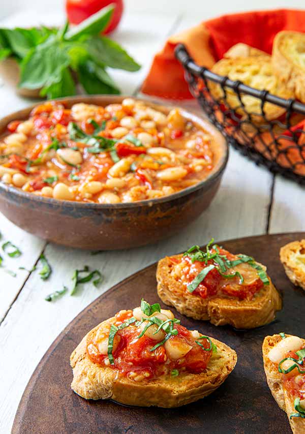 Roasted Tomato and White Bean Crostini on a round wooden board with a bowl of the bean topping on the side and a basket of toasted baguette slices in the background