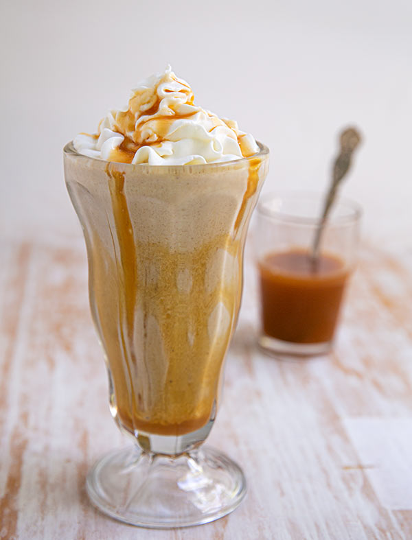 Closeup of SunButter and Caramel Milkshake topped with whipped cream with a cup of caramel sauce in the background on a beige and white table